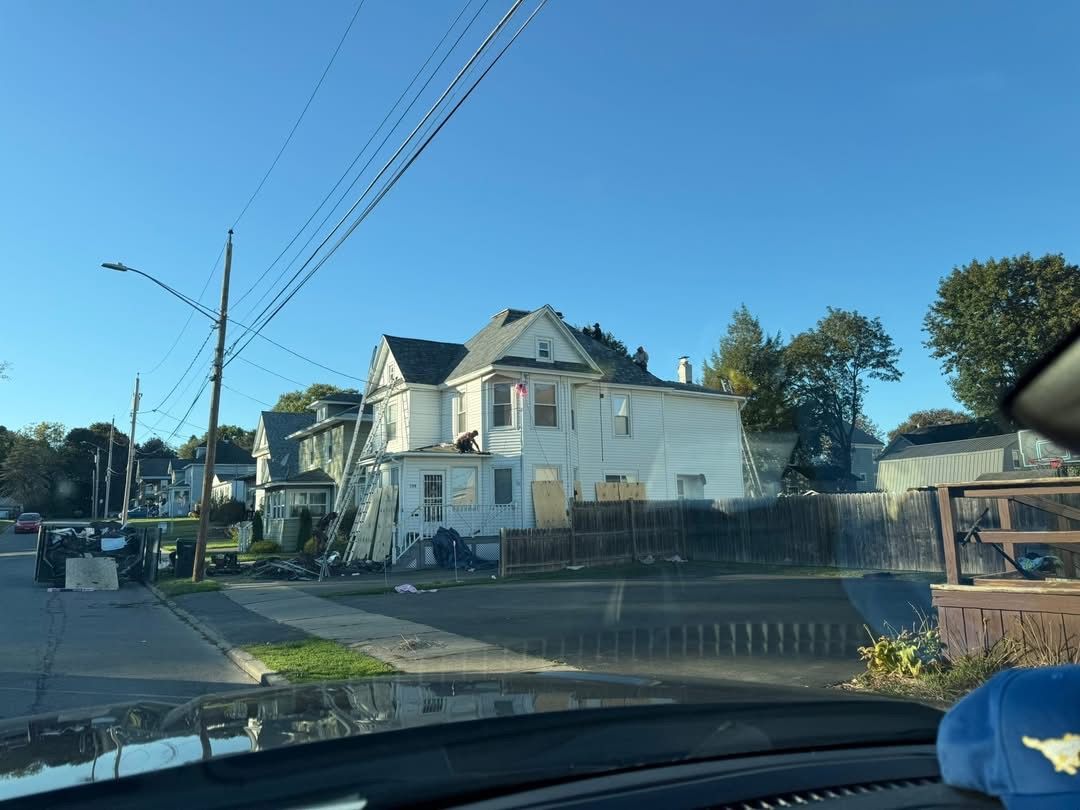 White house with gray roof on a sunny street, utility poles, trees, and blue sky.