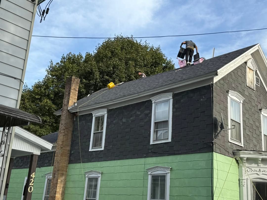 People working on a house roof. Dark gray roof, green and black siding. Cloudy sky.