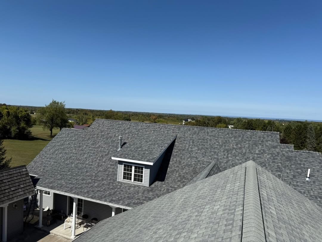 Gray shingled roof of a house on a sunny day. Clear blue sky, green landscape in the distance.