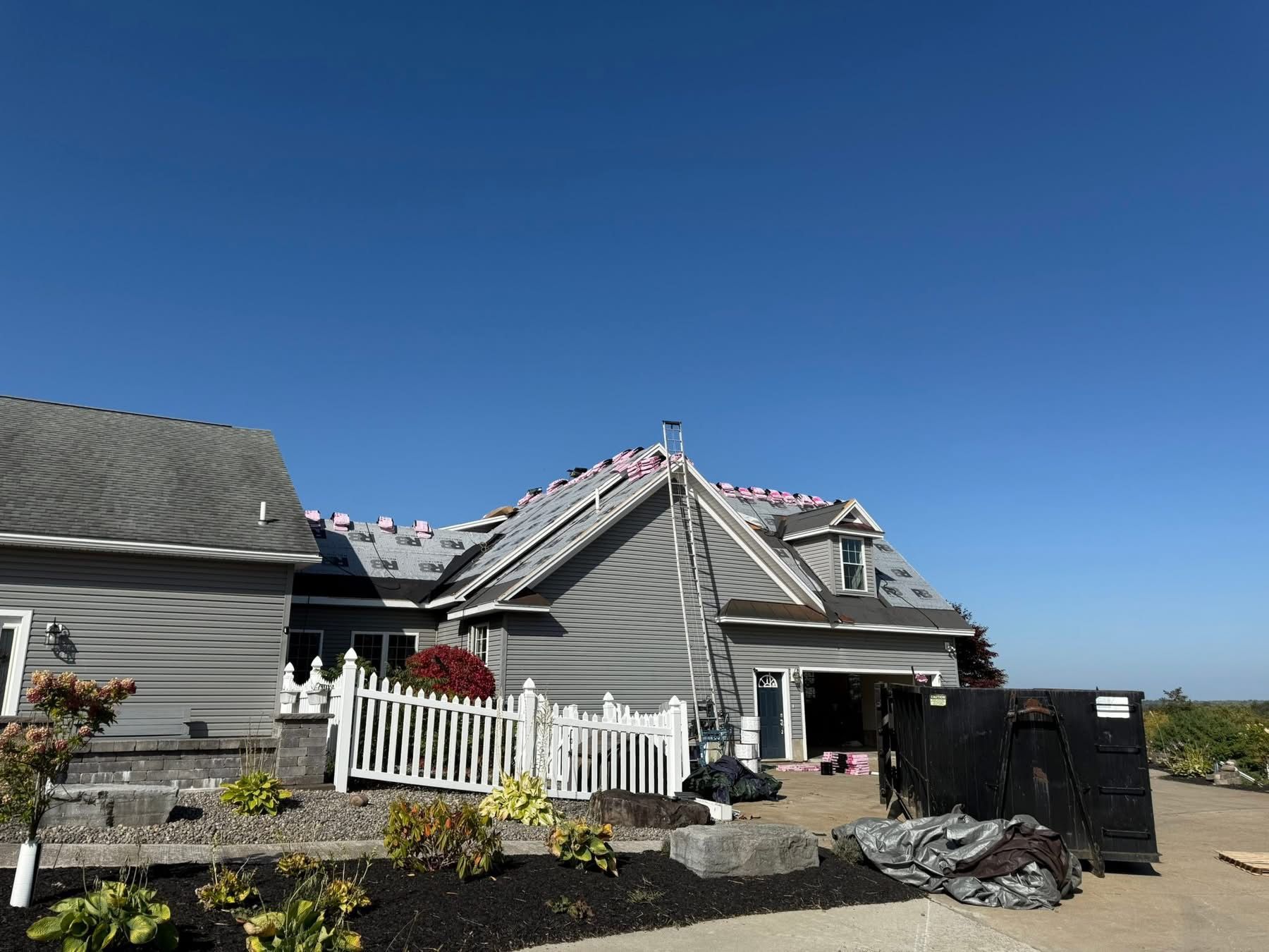 House with roof partially stripped, ladder, and dumpster under a clear blue sky.