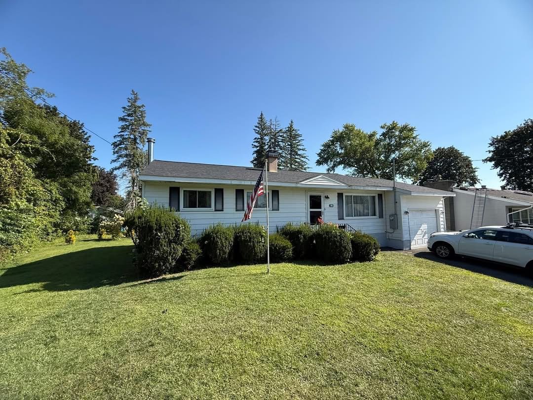 White ranch house with American flag on a sunny day. Green lawn, trees, and parked car are visible.