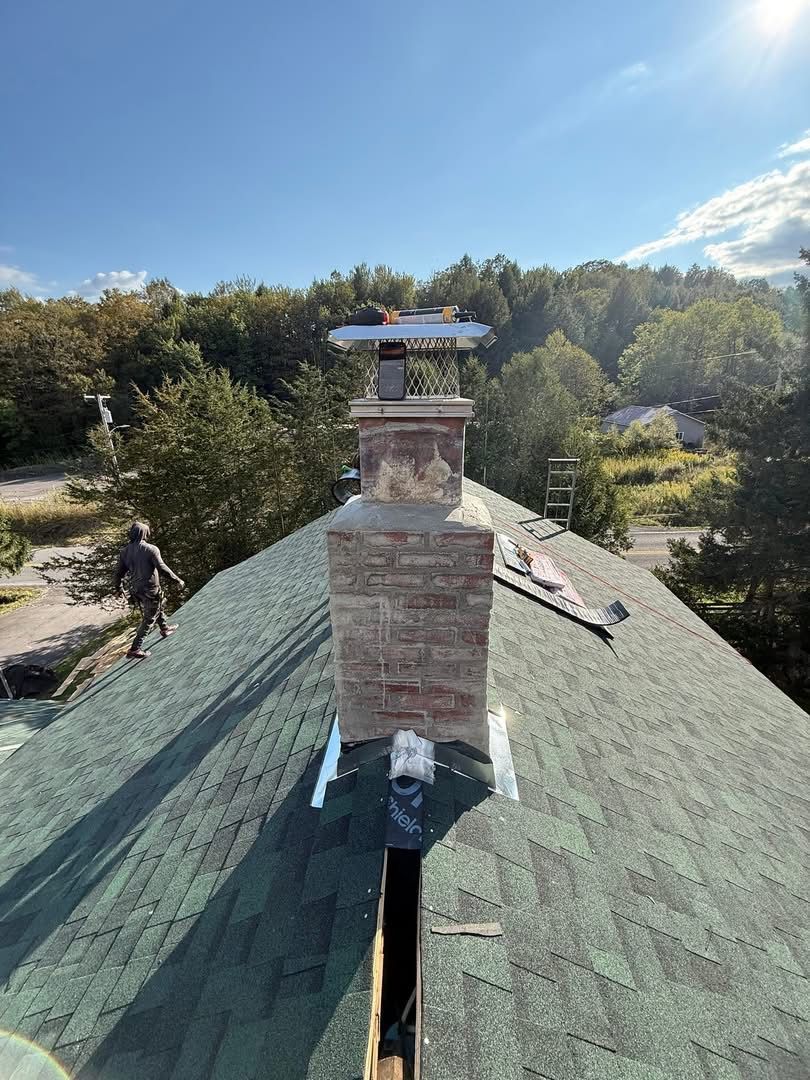 Brick chimney on a green shingled roof with a person working on the side against a backdrop of trees.