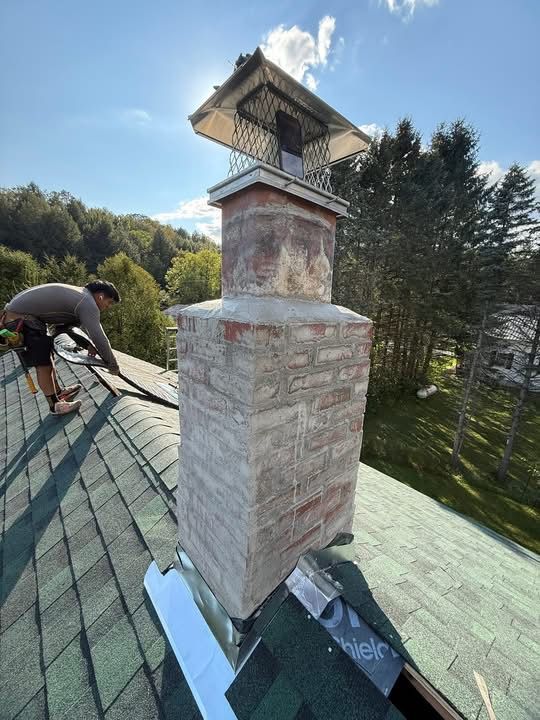 A person repairs a brick chimney on a green shingled roof, set against a backdrop of trees and blue sky.