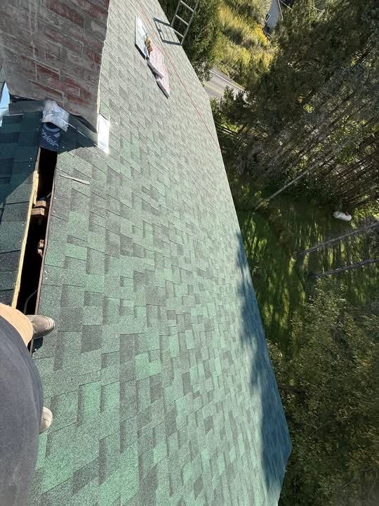 Overhead view of a roof being repaired. Green shingles, exposed underlayment, and a worker's foot are visible.