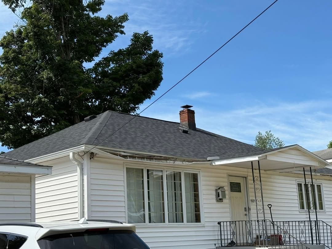 White house with dark gray roof, chimney, and porch, against a blue sky with tree.