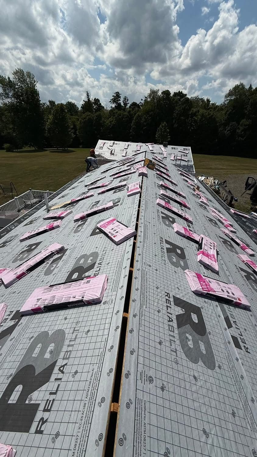 Rooftop with black underlayment, pink insulation, and exposed wooden beams under a partly cloudy sky.