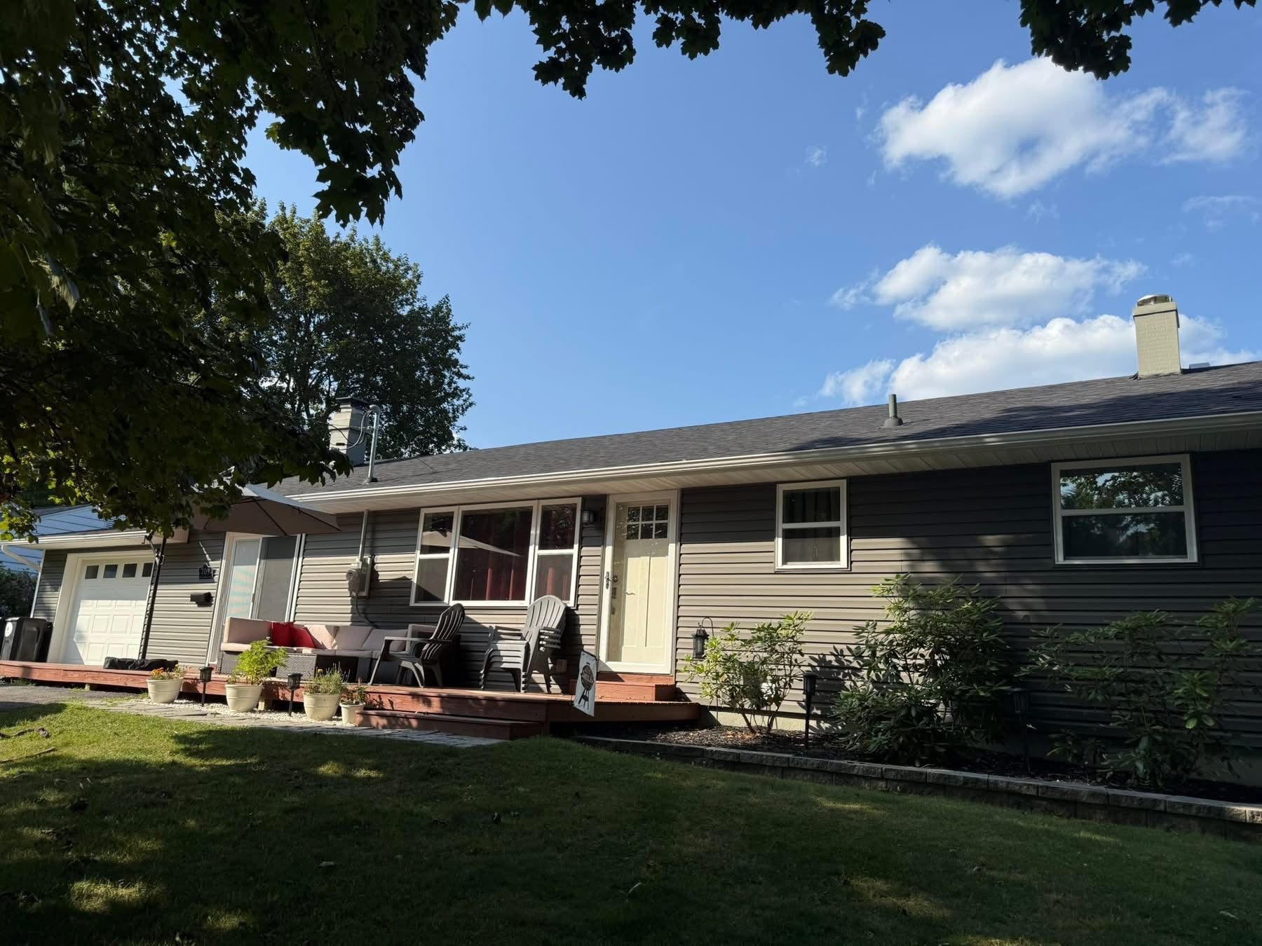 Back view of a house with gray siding, a deck, and green grass under a blue sky with clouds.