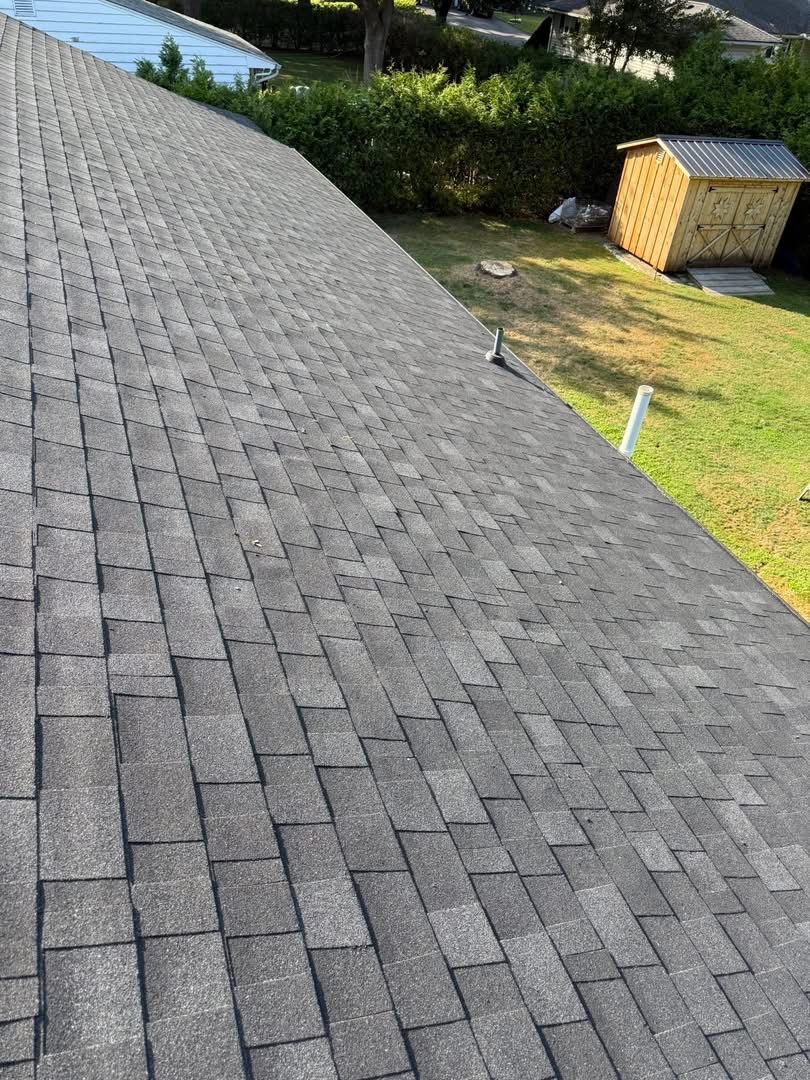 Close-up of a gray shingled roof with a shed and grassy yard in the background.