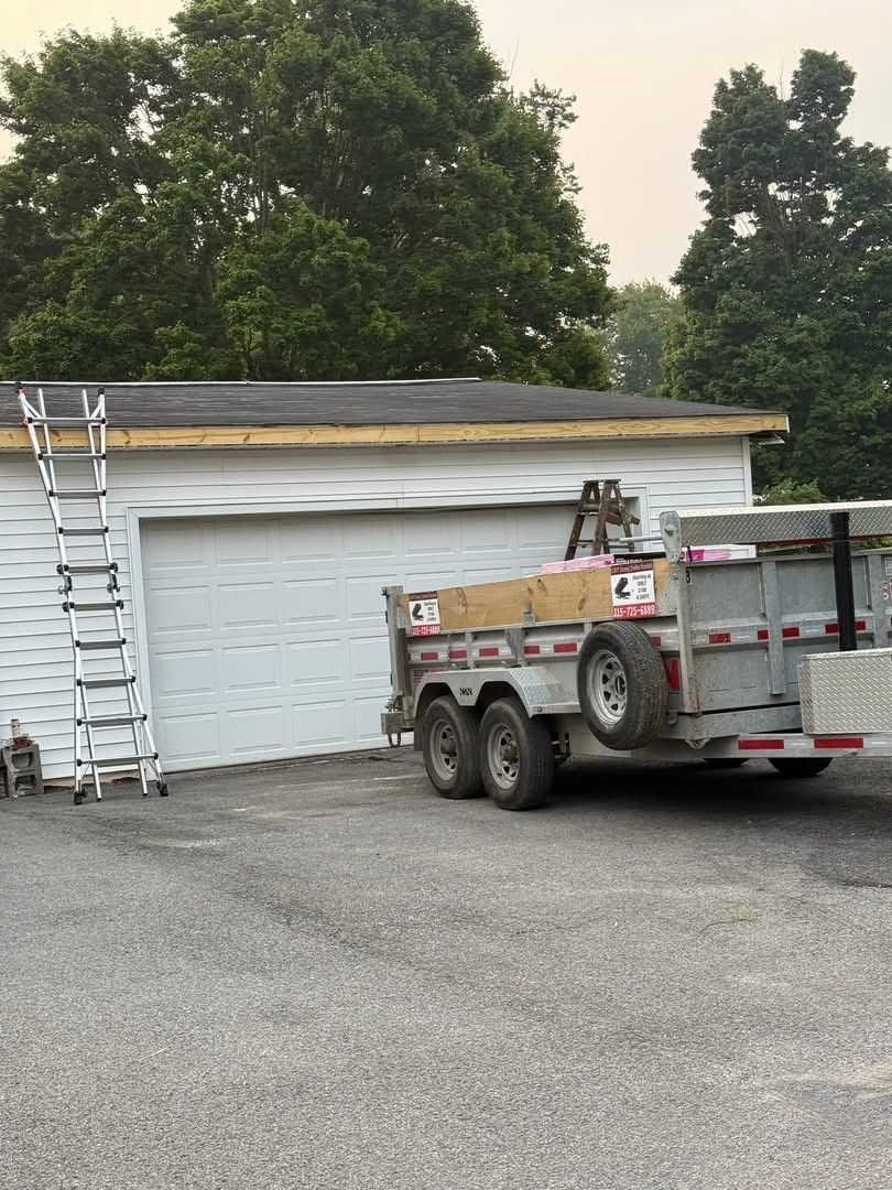 White garage with ladder and trailer for roof work. Trees in background.