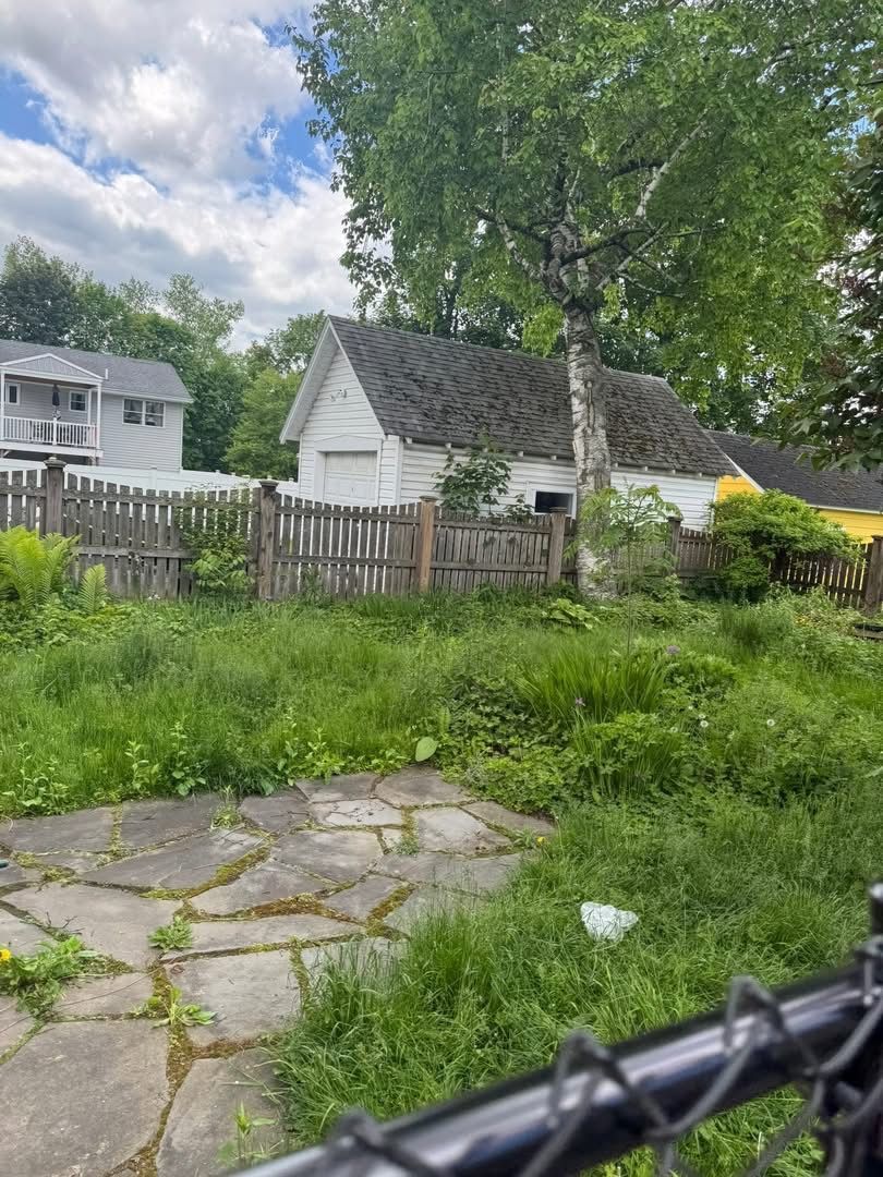 Overgrown yard with a dilapidated white garage and fence, overcast sky.