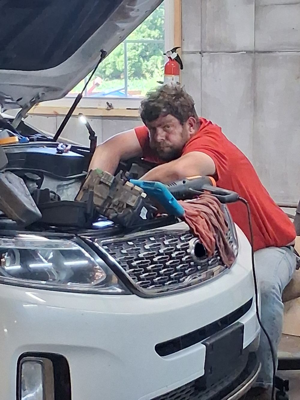Mechanic in a red shirt working under a raised car hood in a garage | Kensinger Repair Center