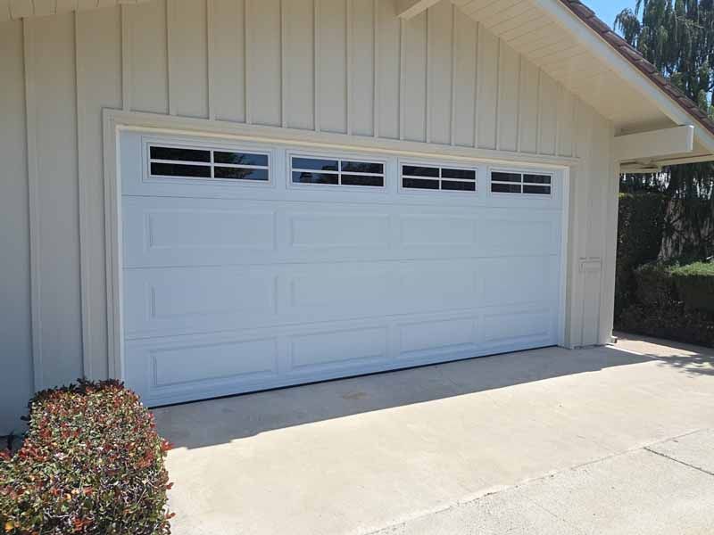 White two-car garage door on a beige house with a sunlit driveway and small bush nearby