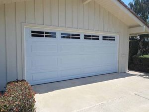 White two-car garage door on a beige house with a sunlit driveway and small bush nearby