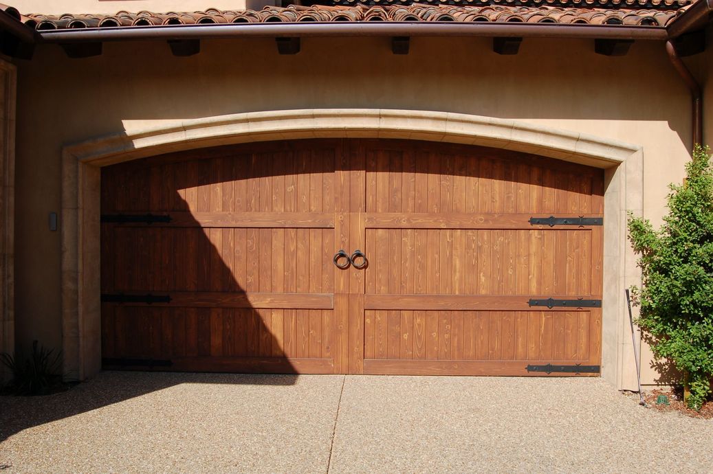 Wooden double garage door with arched trim on a beige house exterior, partly shaded by sunlight
