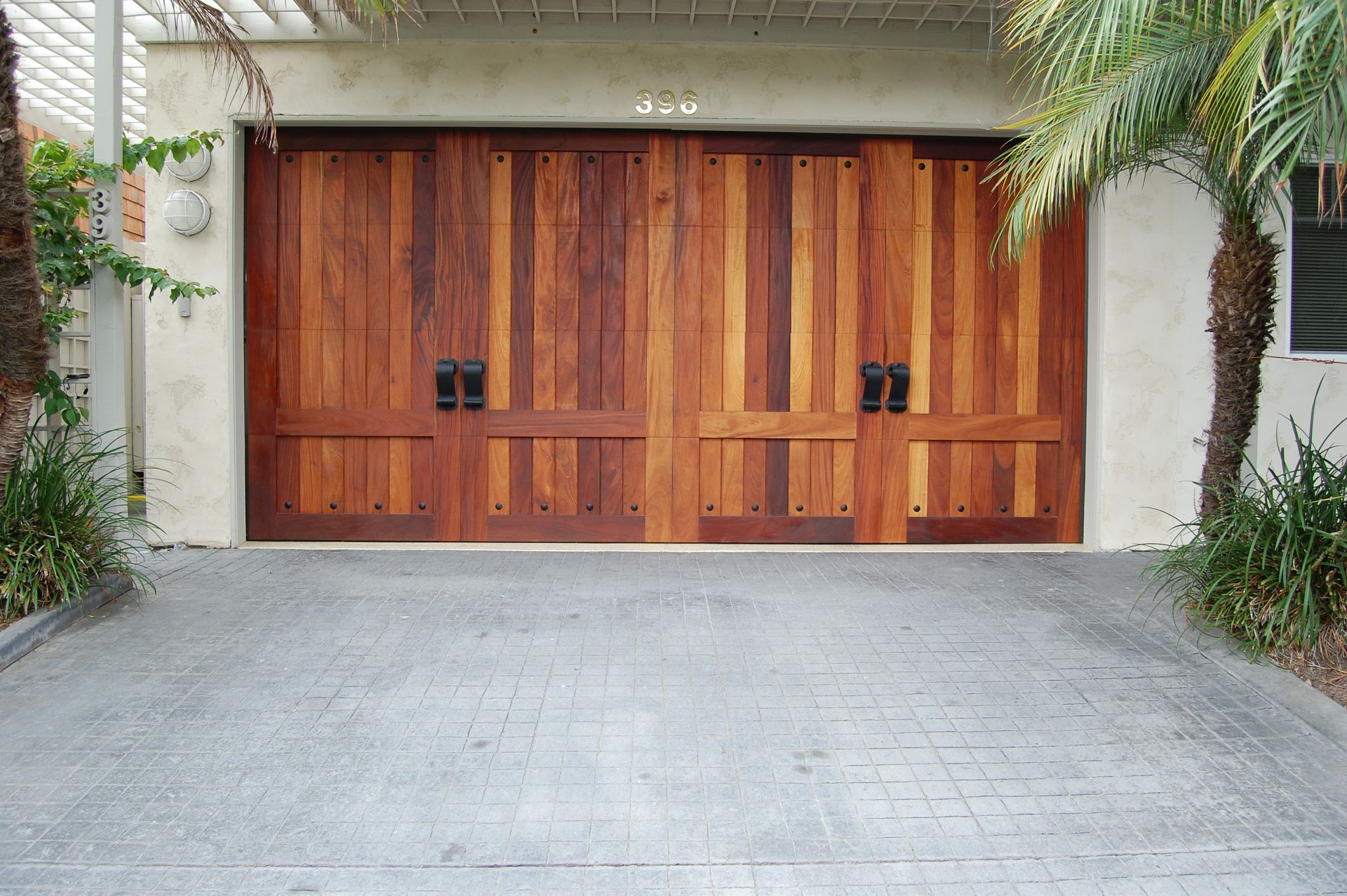 Wooden double garage doors with black handles set in a white stucco wall, framed by plants and a concrete driveway.
