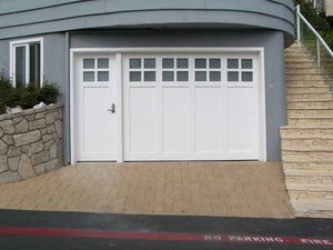 White garage door with side entry door beside stone wall and stairs on a sloped driveway
