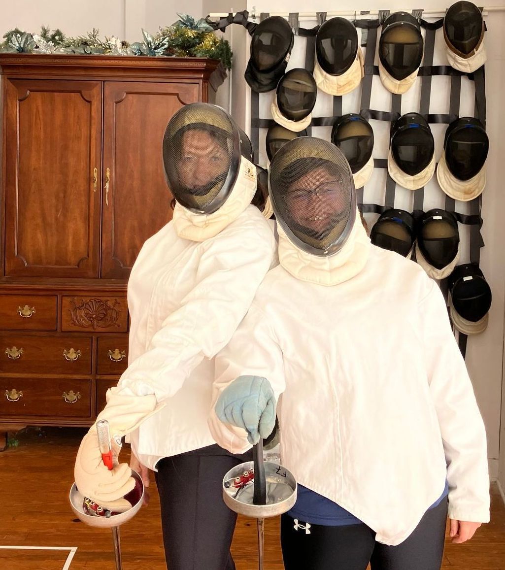 Two women wearing fencing gear smile through mesh masks