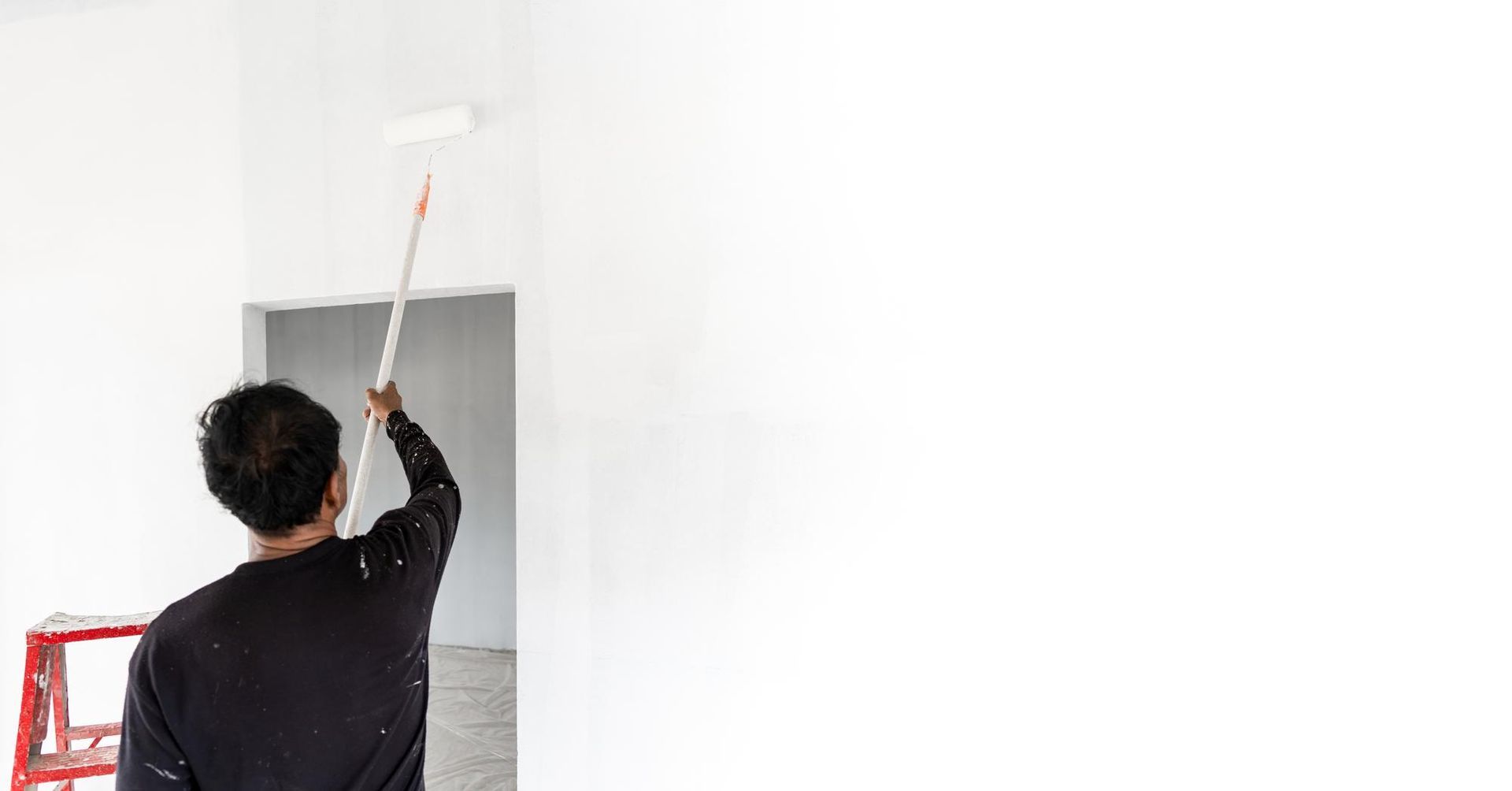 Person painting a white wall with a roller, standing near a red ladder in a room.