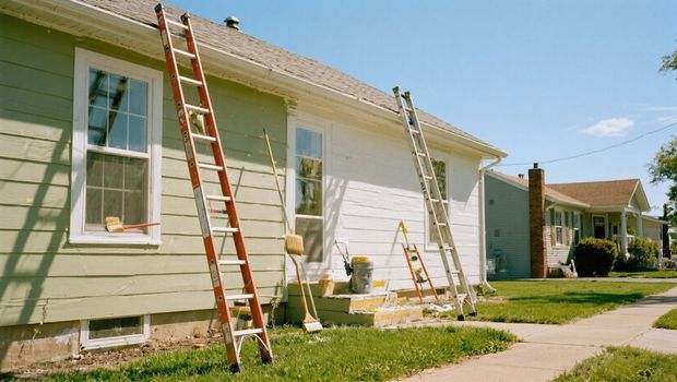 Two ladders lean against a house being painted; green and white sections. Sunny day.