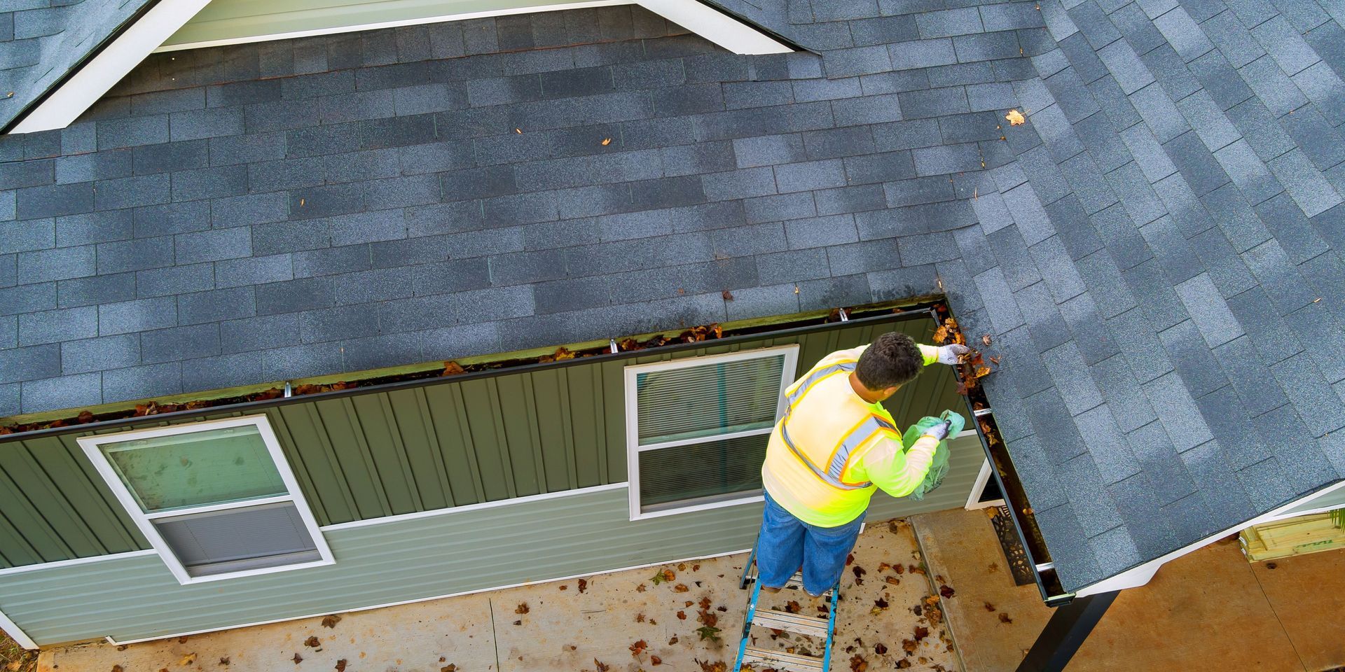 Person on a ladder cleaning leaves from a house gutter, wearing a safety vest and gloves.