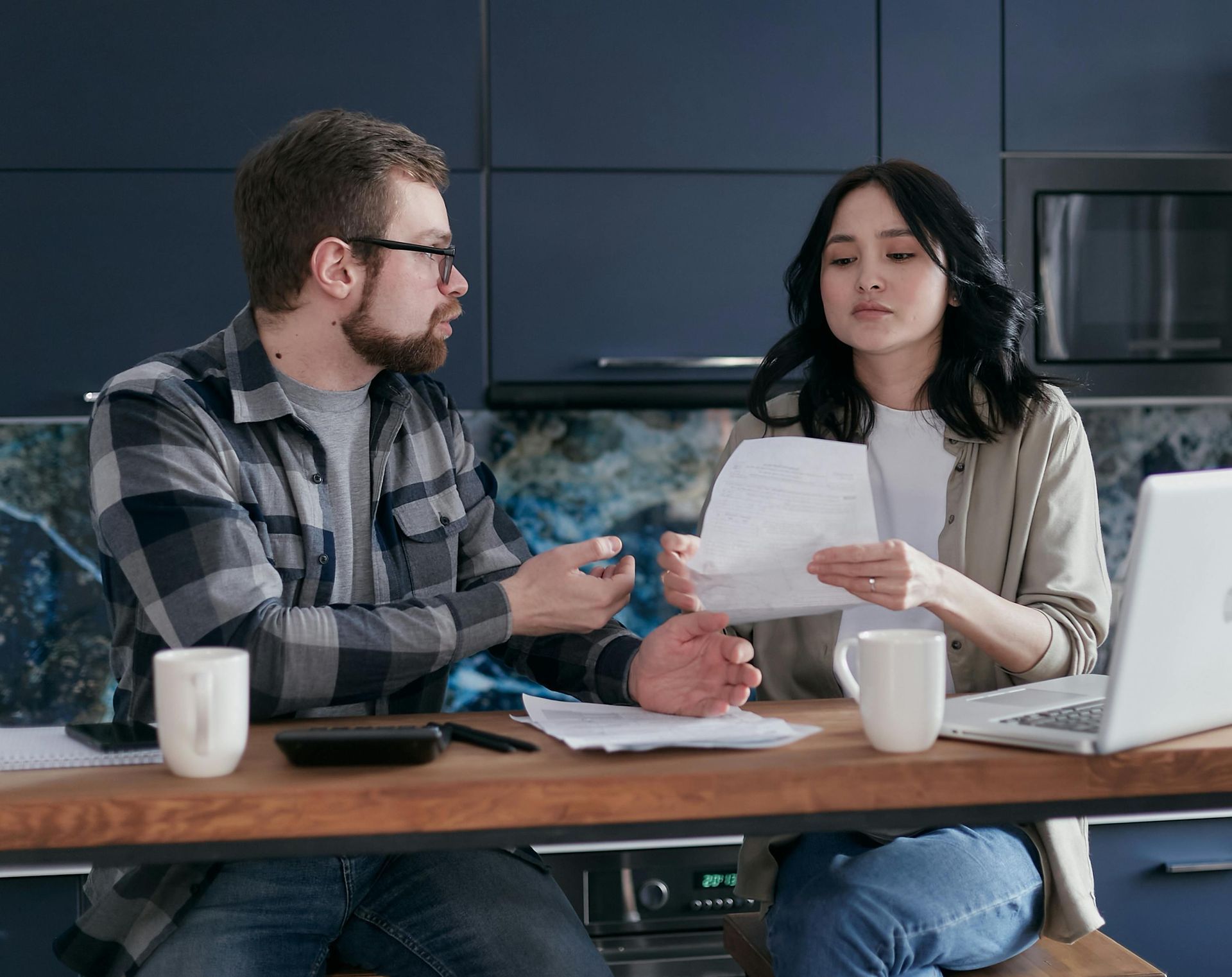 Couple in kitchen, reviewing documents with serious expressions, calculator and laptop on table.