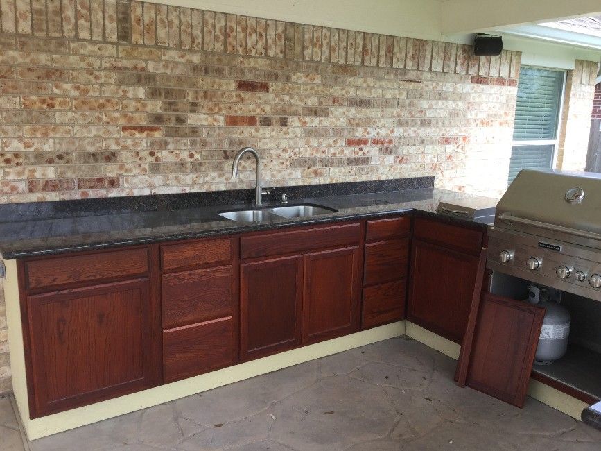 Outdoor kitchen with brick wall, granite countertop, brown cabinets, and a grill.