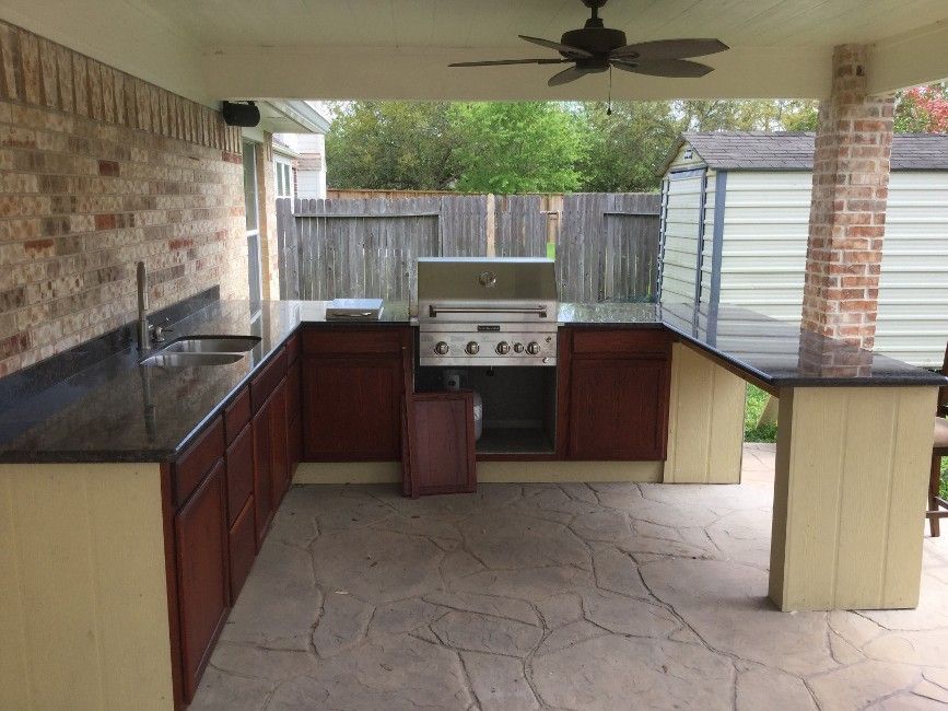 Outdoor kitchen with built-in grill, cabinets, sink, and bar under a covered patio with brick and stone accents.