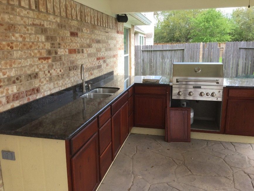 Outdoor kitchen with brick wall, granite countertop, sink, cabinets, and grill.