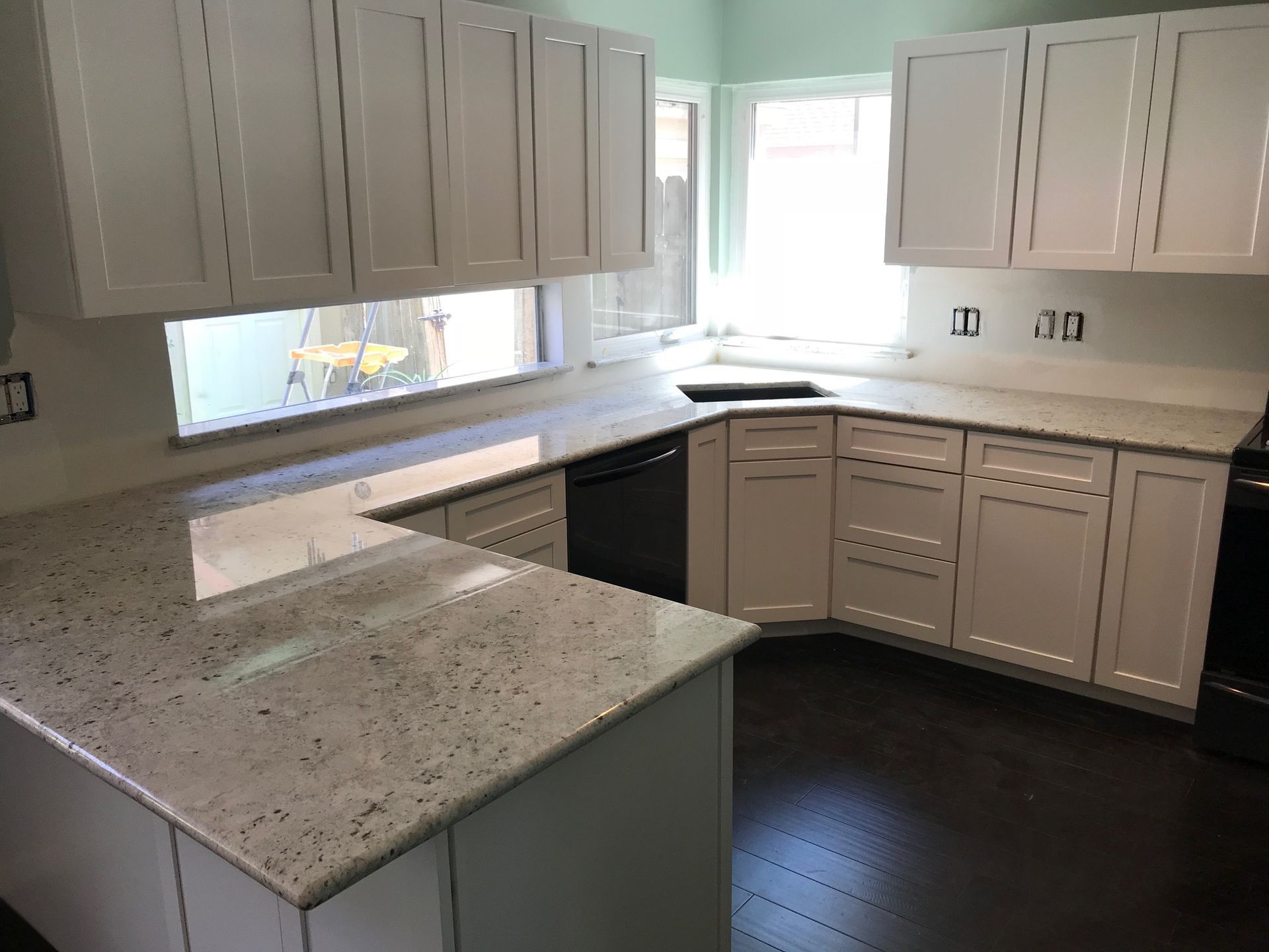 Empty kitchen with white cabinets, speckled countertops, and dark wood flooring near a bright window.
