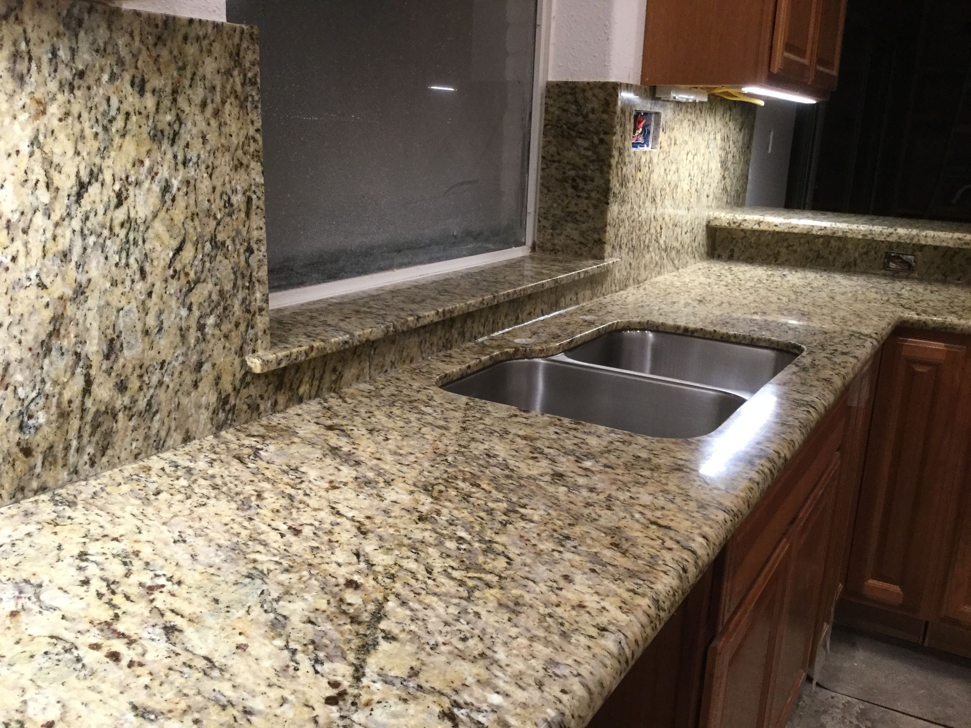 Granite countertop with a double sink, beside a window and wooden cabinets, lit from above.