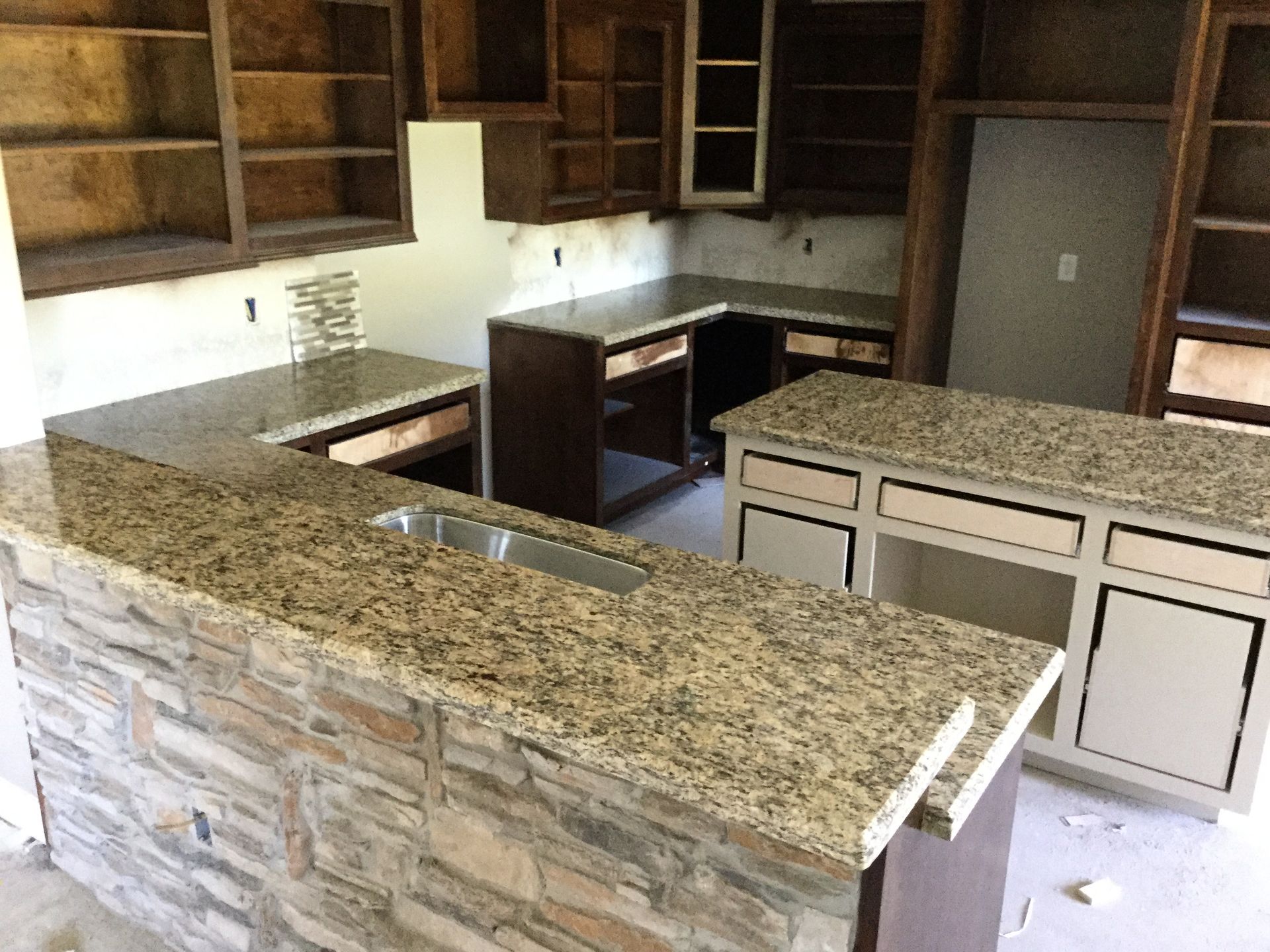 Kitchen under construction with granite countertops, wooden cabinets, and a stone facade on island.