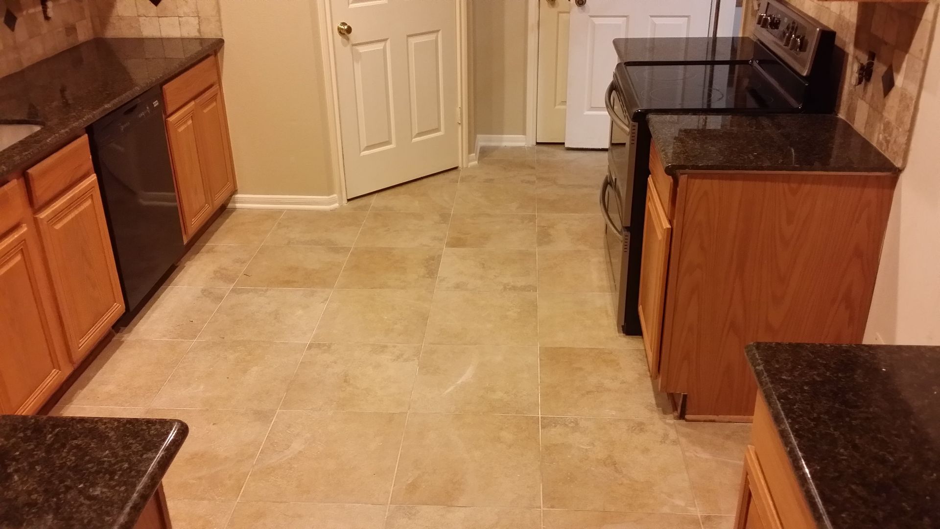 Kitchen with brown cabinets, black countertops, and beige tiled floor. A stove and dishwasher are present.