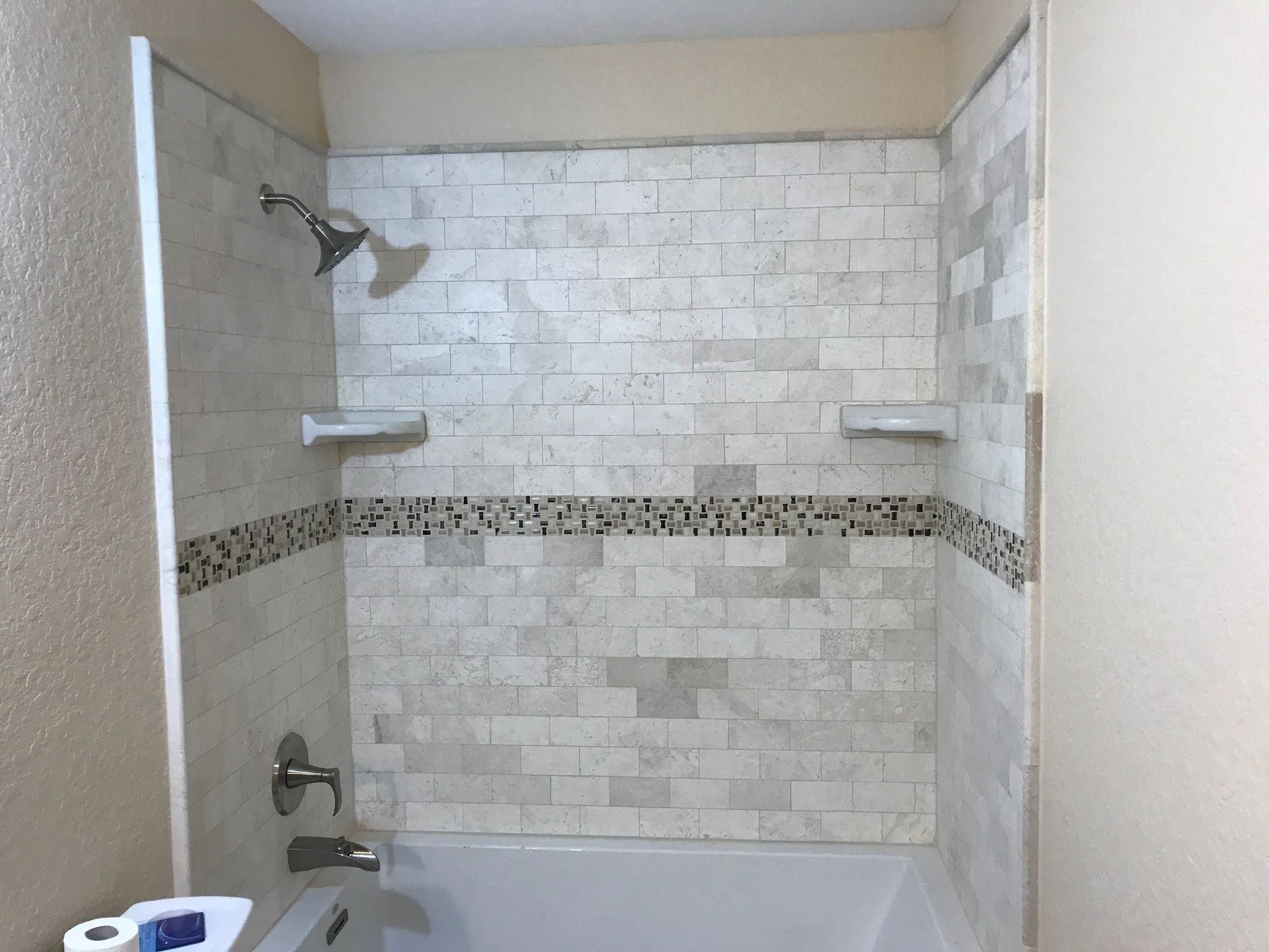 Bathtub and shower with light stone tile walls, small mosaic accent, and showerhead.