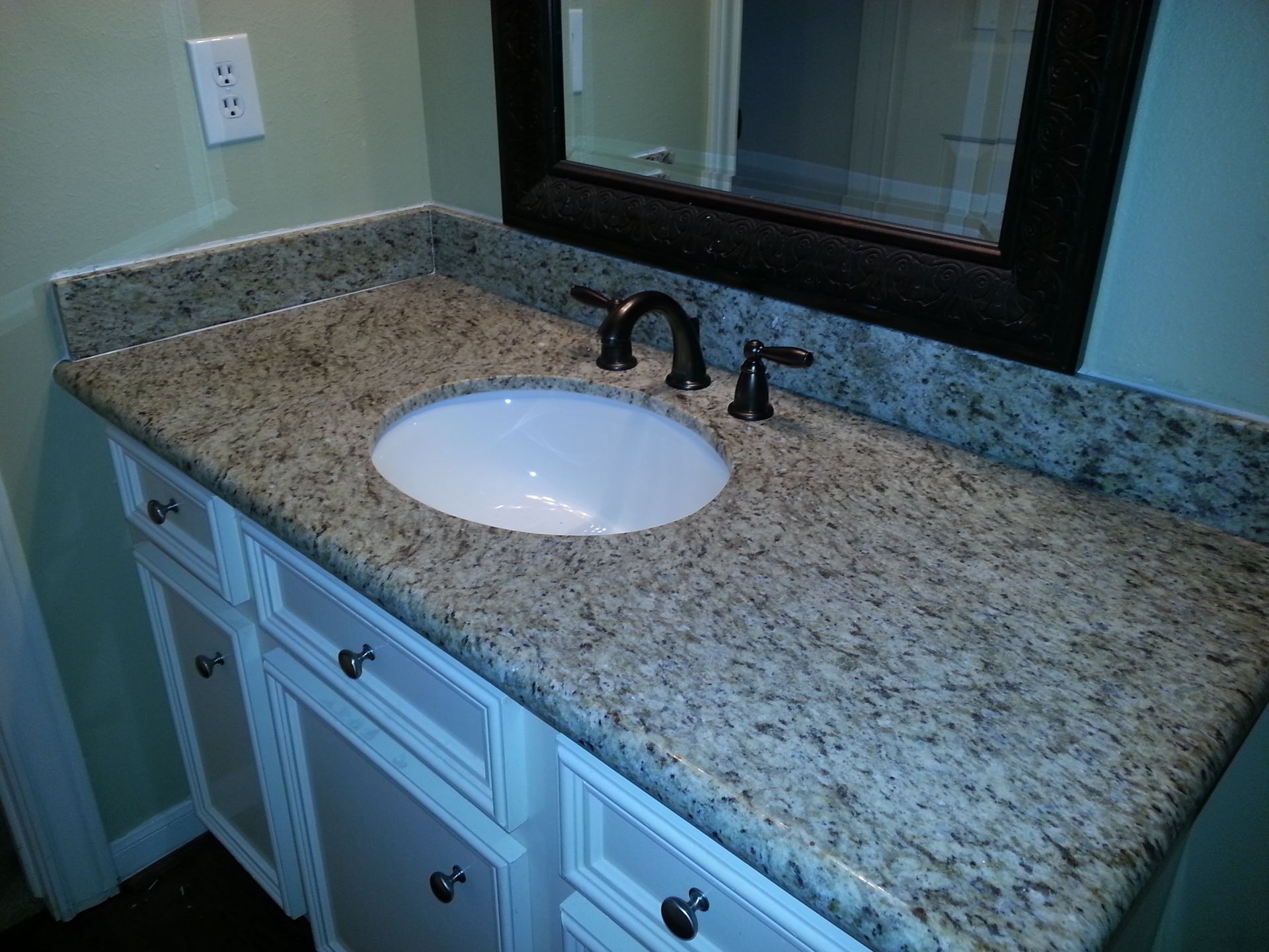 Bathroom vanity with granite countertop, oval sink, black faucet, white cabinets, and mirror.