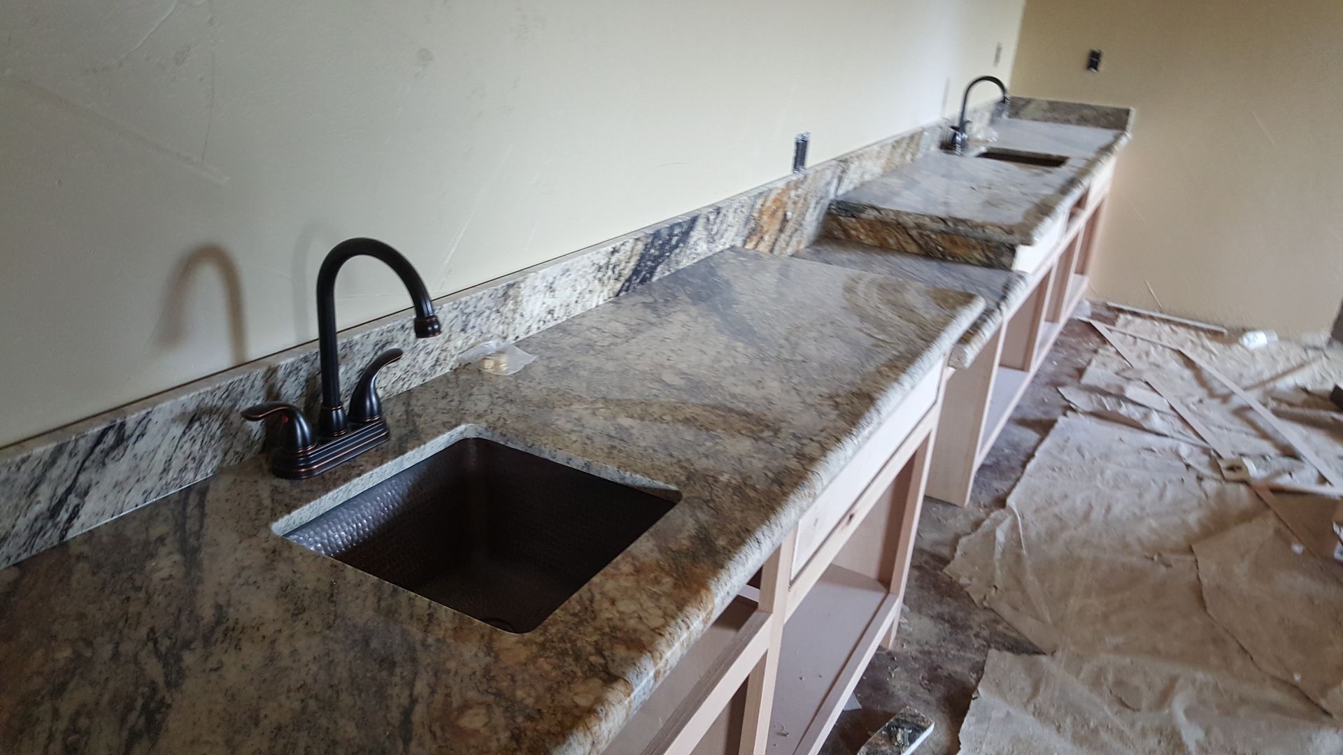 Granite countertop with sinks, installed in unfinished wooden cabinets. Dark faucets and beige walls.