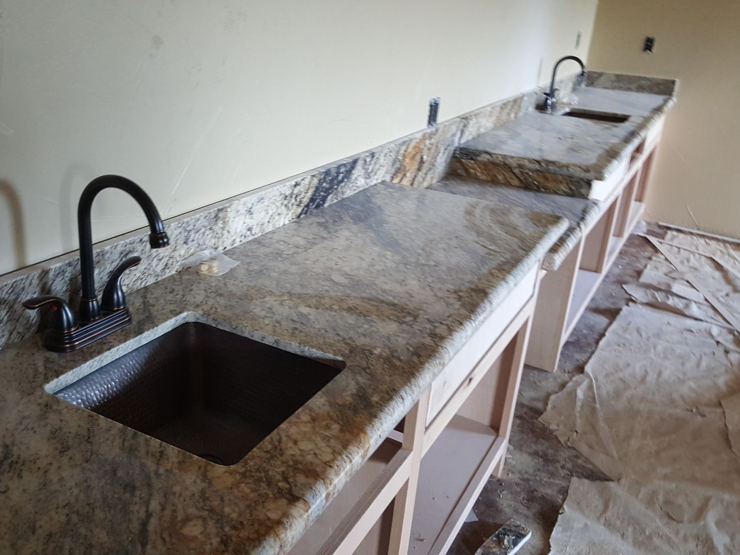 Two installed granite bathroom vanities with black faucets, stone backsplashes, and unfinished wooden cabinets.