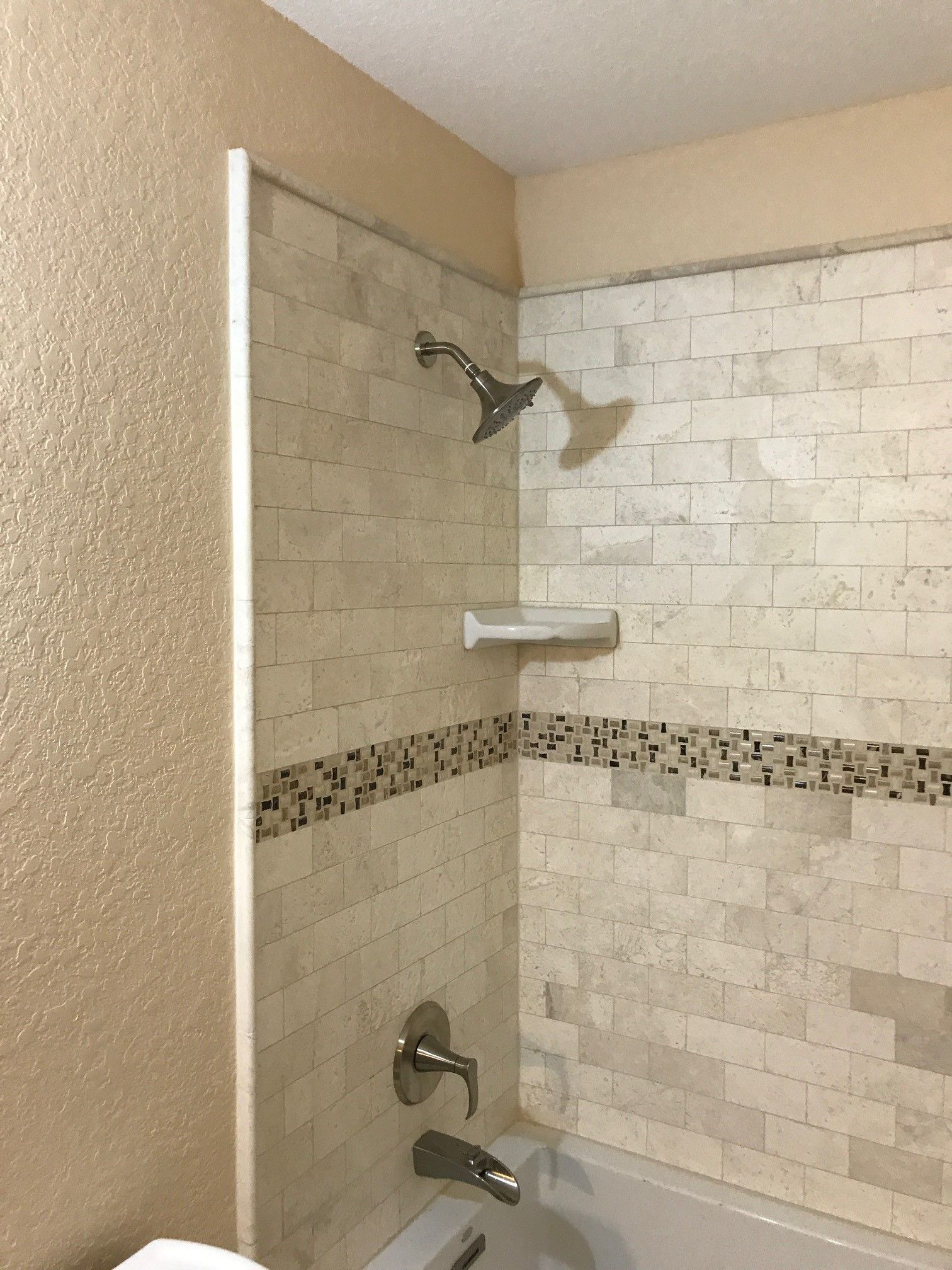 Shower stall with beige tile, showerhead, shelf, and faucet. Beige and mosaic tile accents.