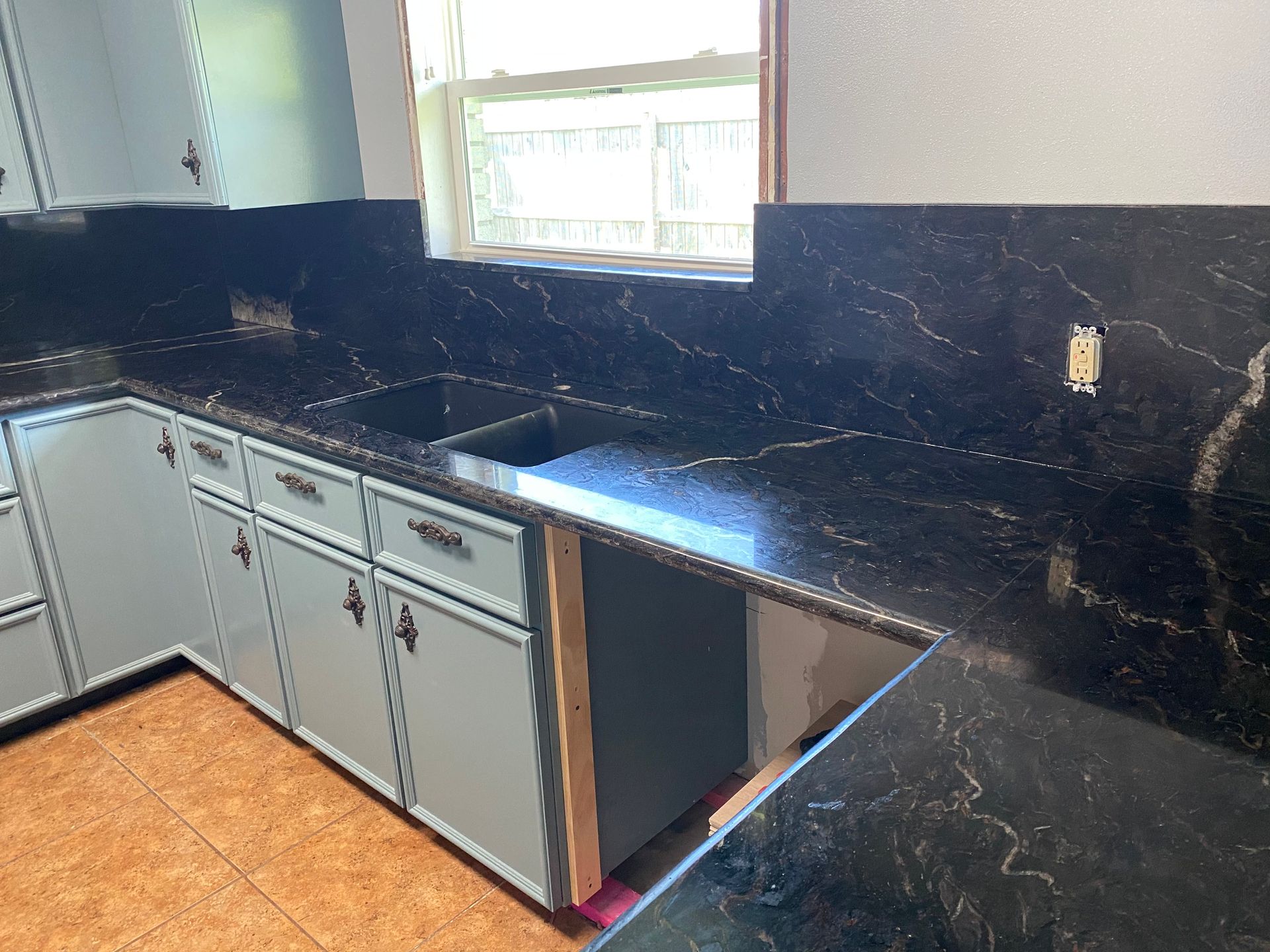 Kitchen with black marble-like countertops, light blue cabinets, a window, and a sink.