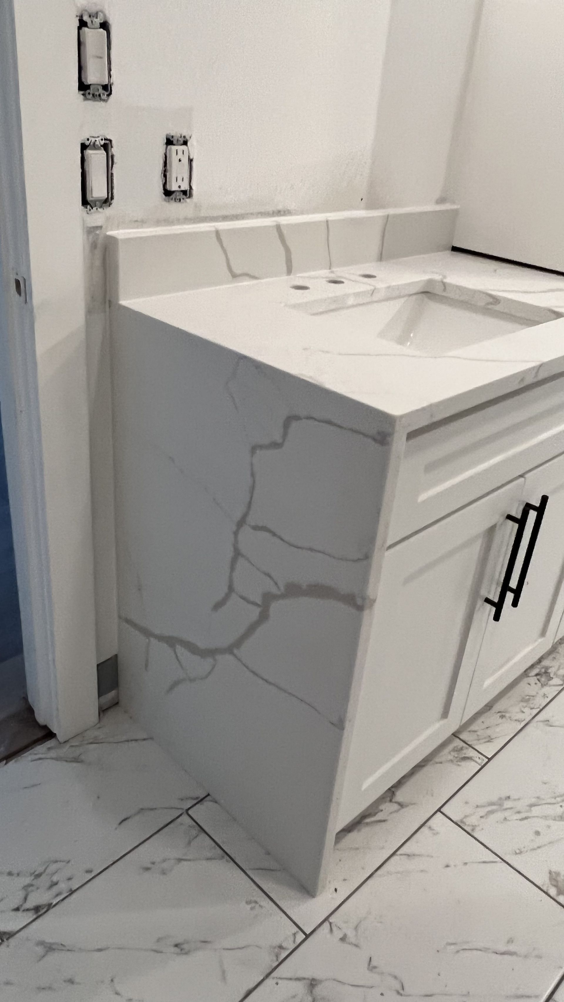 White bathroom vanity with marble countertop and cabinet. Black hardware, unfinished wall, and tile floor.
