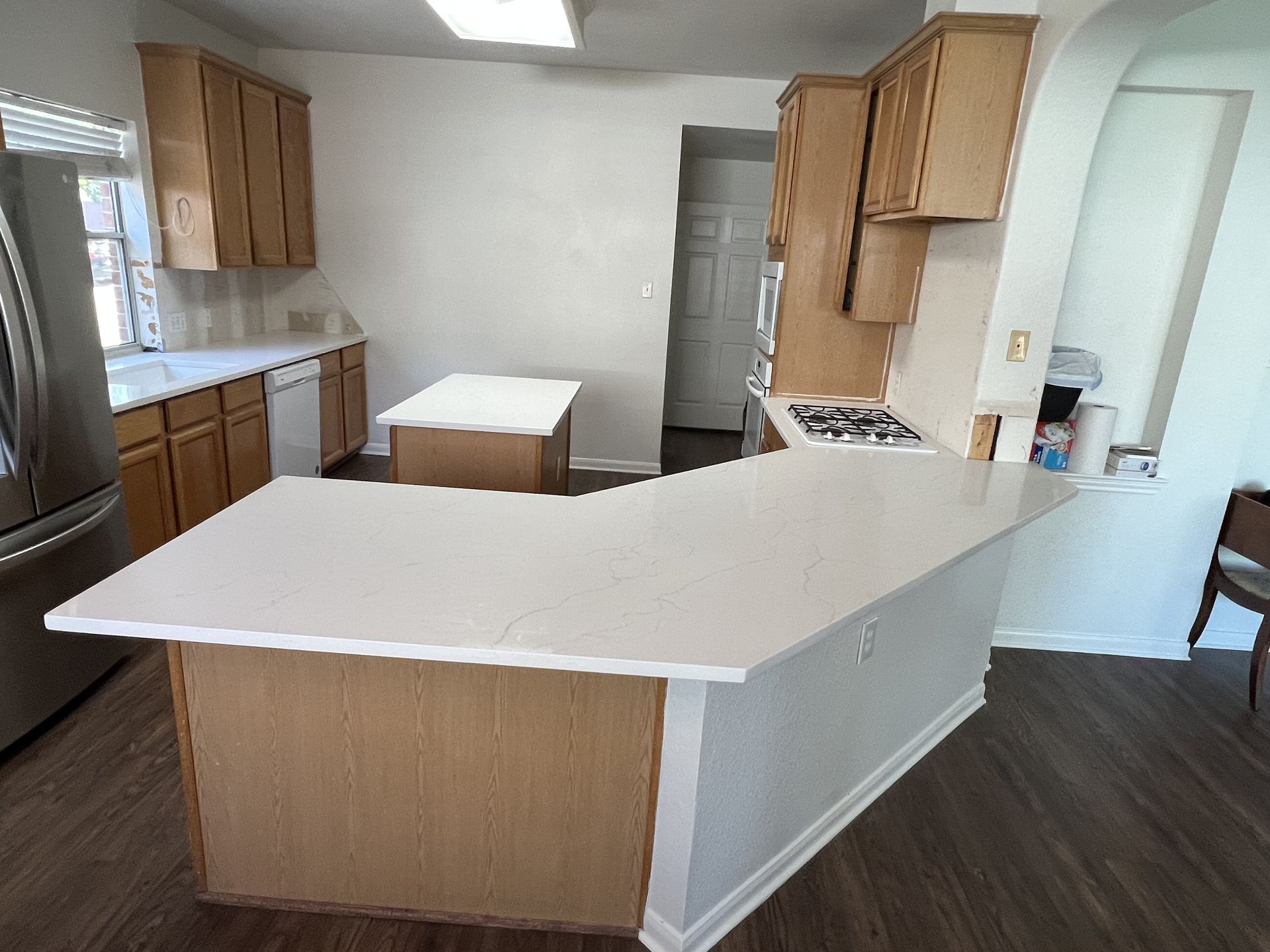Empty kitchen with white countertops, wood cabinets, stainless fridge, and dark wood flooring.