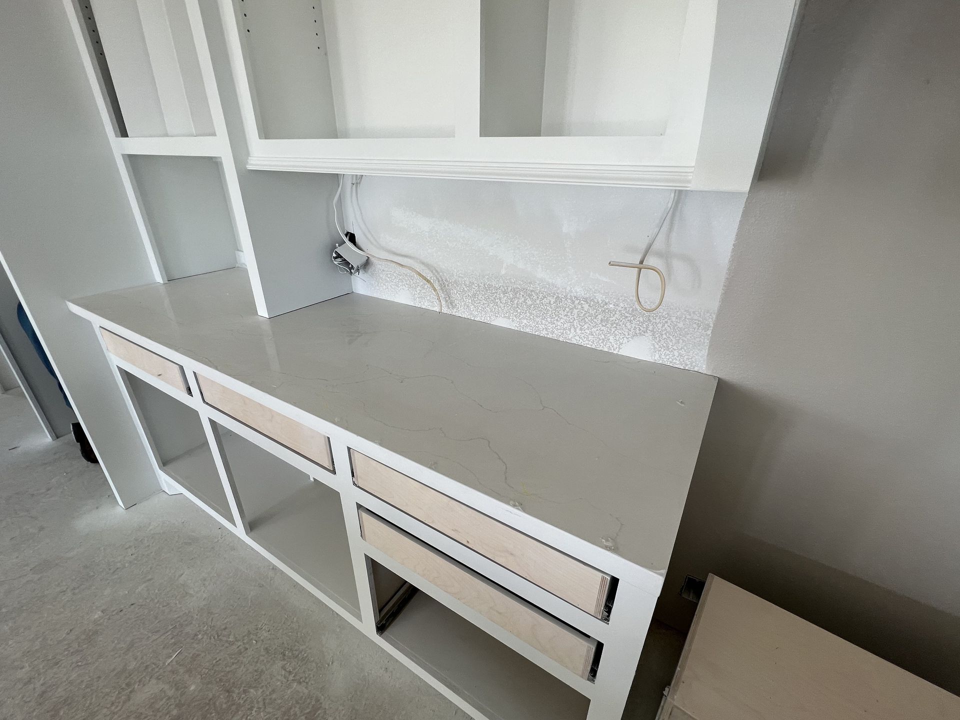 White built-in desk with drawers and open shelves in a carpeted room, set against a light wall.