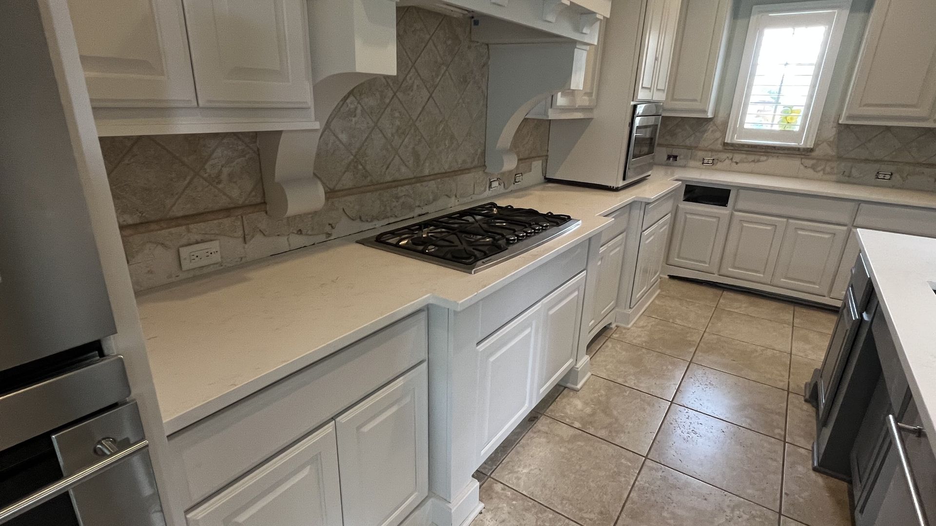 White kitchen with cabinets, countertops, and appliances. Gas cooktop, tile backsplash, and window.