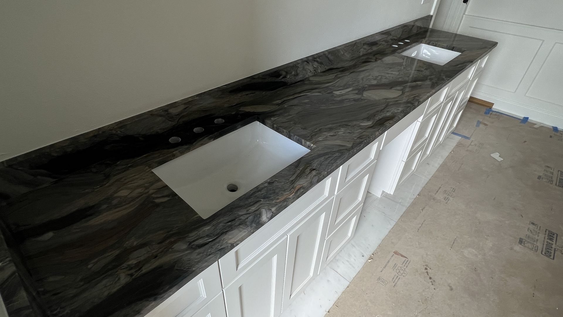 Long granite countertop with two white sinks on white cabinets in a room under construction.