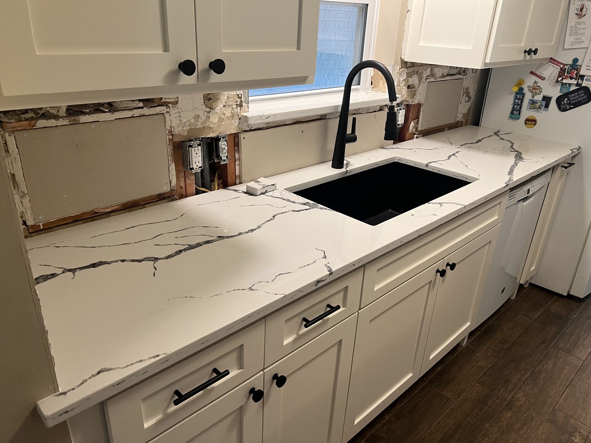 Kitchen with damaged wall above marble countertop, sink, and white cabinets; wall missing near backsplash and plumbing exposed