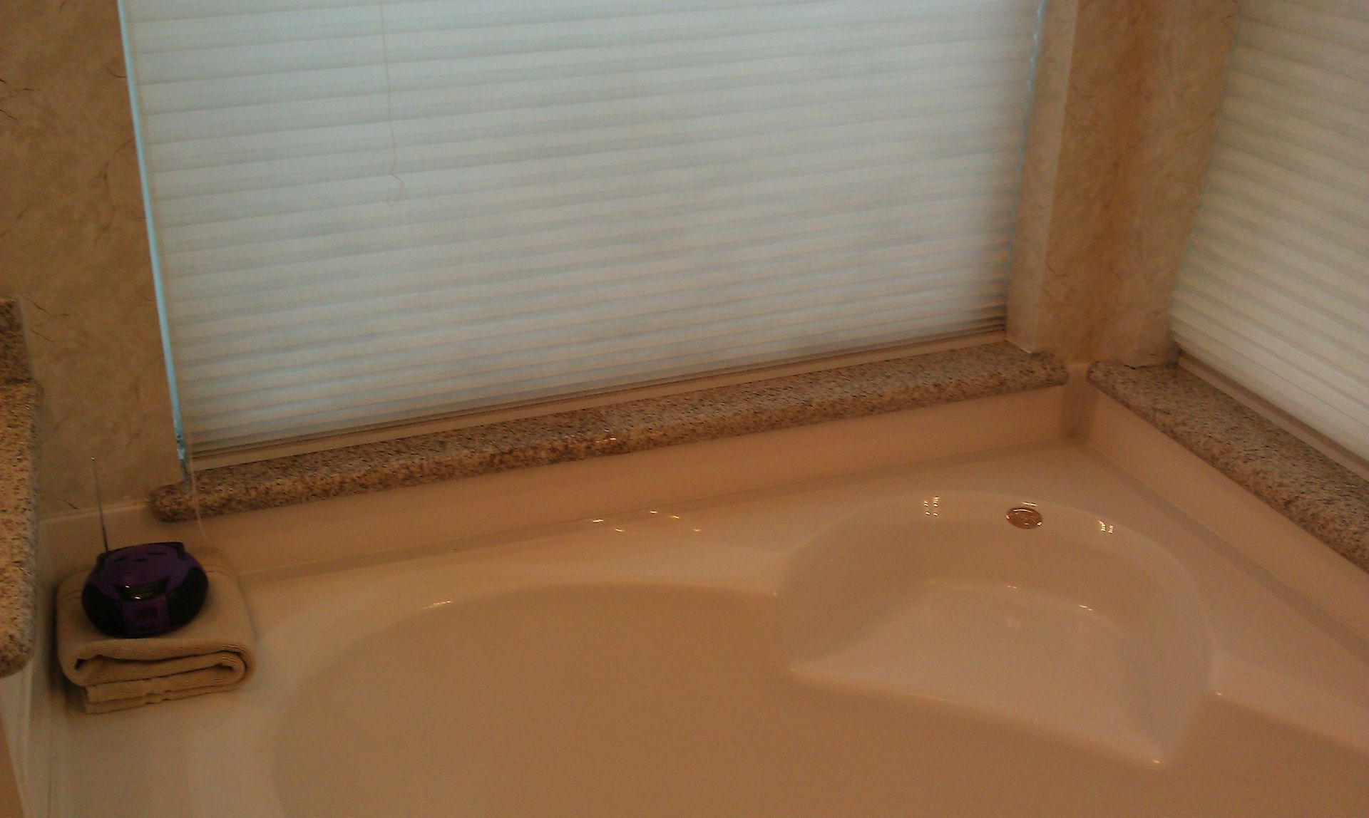 Bathtub with corner marble trim and a closed window blind. A towel and candle are on the corner.