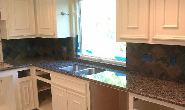 Kitchen with granite countertops, cream cabinets, and dark tile backsplash. Window in center.