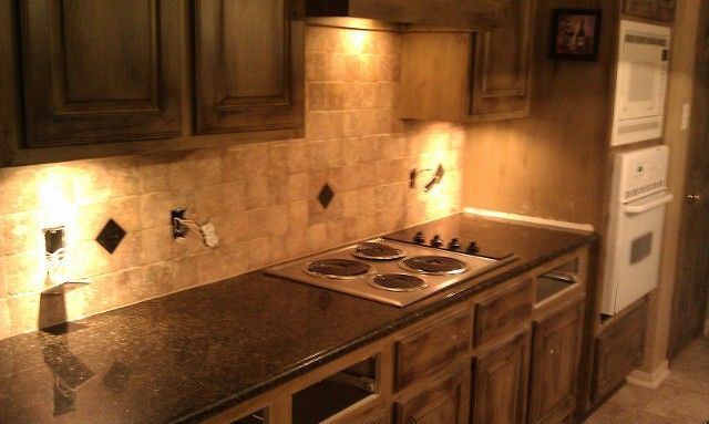 Kitchen with dark cabinets, black countertops, and beige tile backsplash. Stove top in view, lit from above.