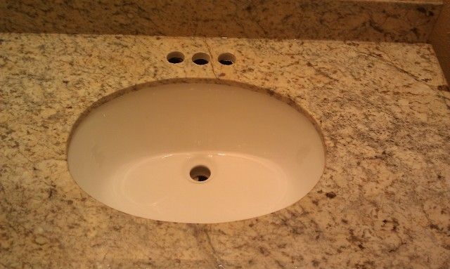 White oval sink set in a speckled tan granite countertop. Three faucet holes are visible.