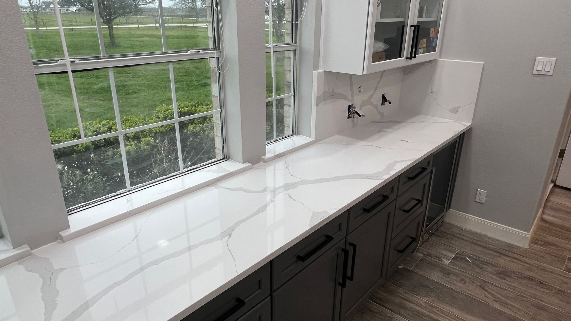 Bright kitchen counter with white quartz top, dark cabinets, and large windows overlooking greenery