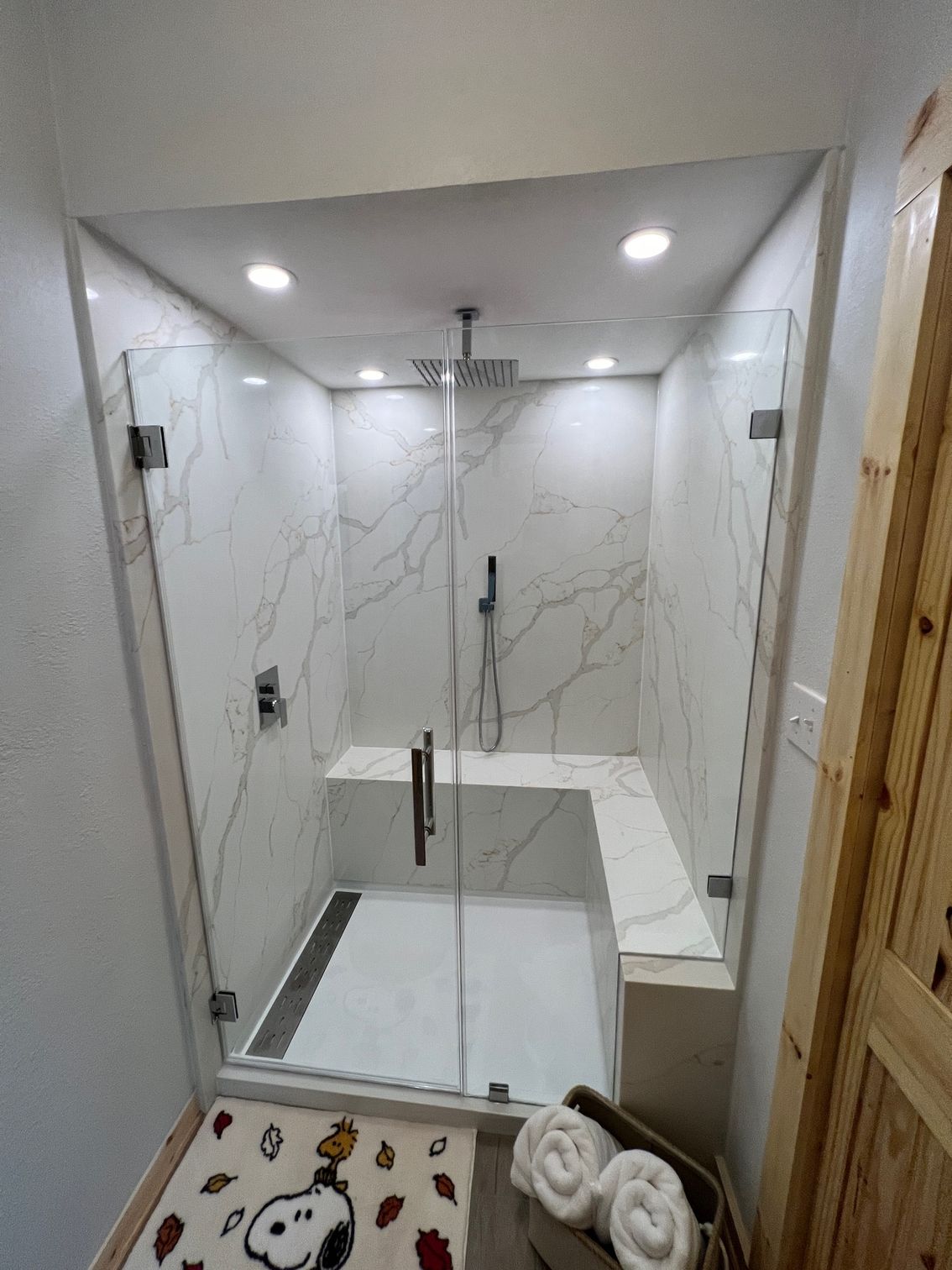 Modern white tiled shower with glass door, bench, recessed lights, and towels on the floor